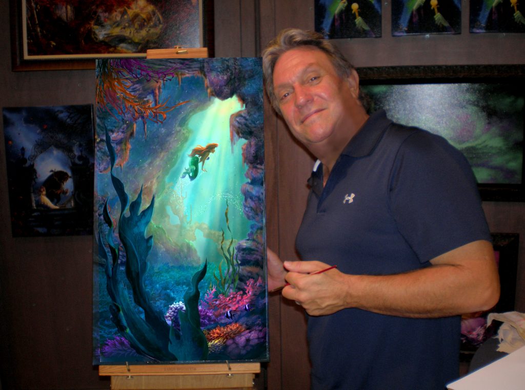 Smiling man in a dark polo posing beside a tall vibrant underwater painting on an easel in a gallery setting.